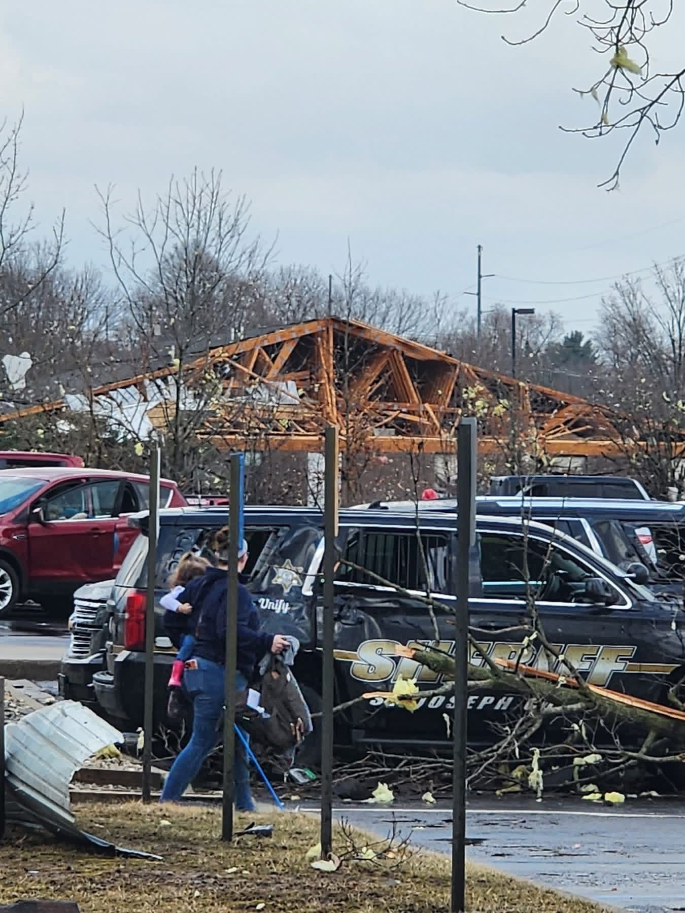 Storm-damaged parking lot with crushed black SUV, debris, and a person walking near parked cars