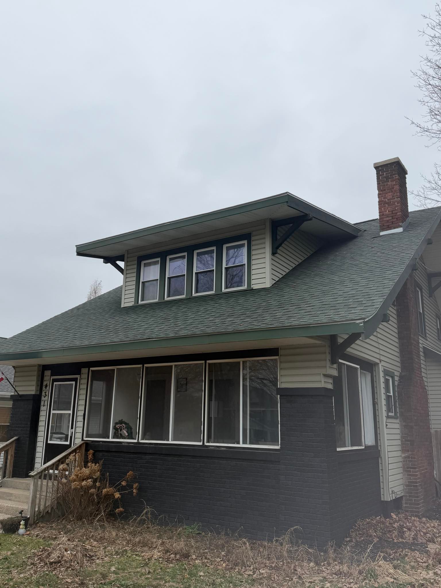 Small house with a green roof and brick chimney under a cloudy sky, surrounded by sparse shrubs.