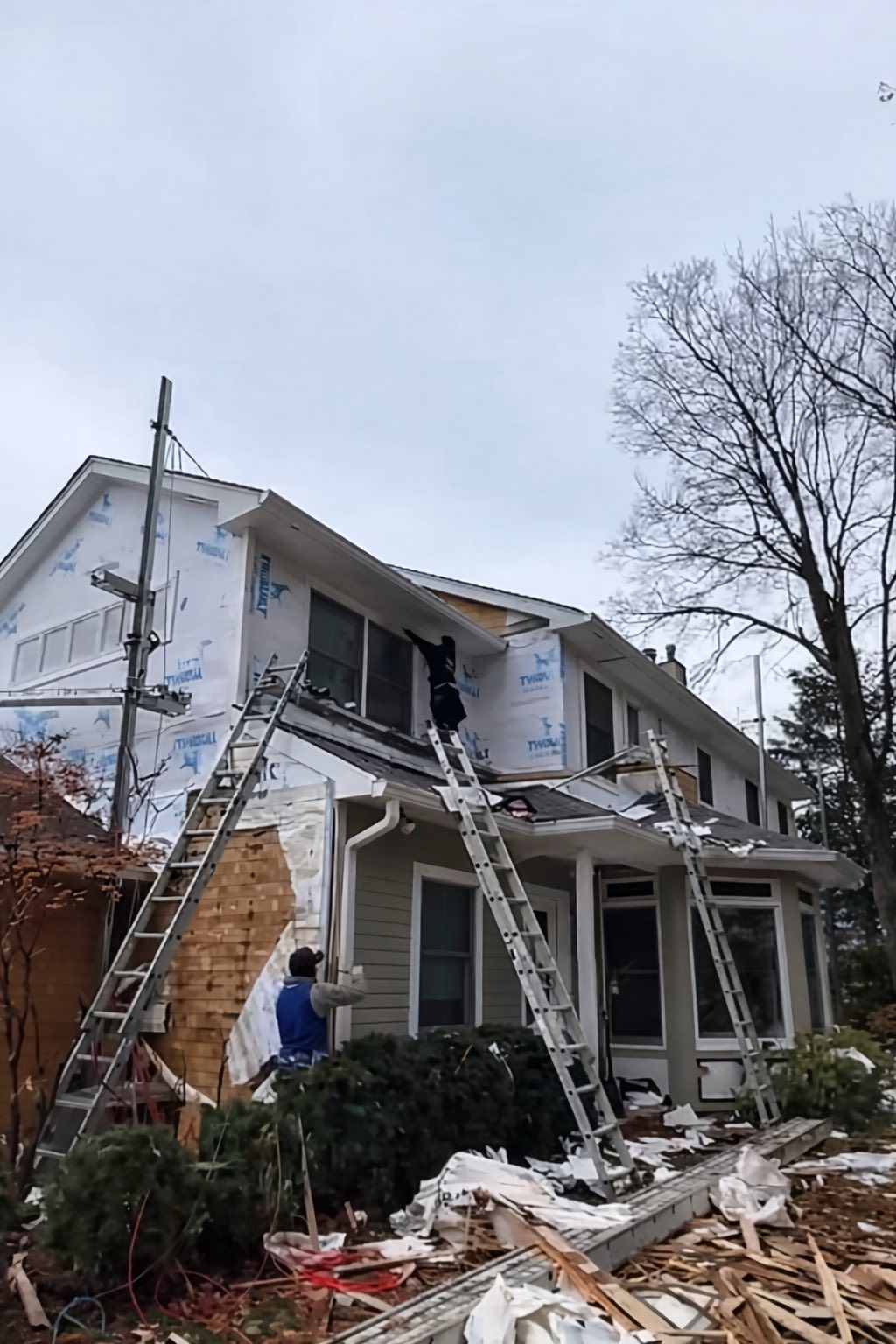 Damaged two-story house with collapsed porch and debris after storm, worker standing outside
