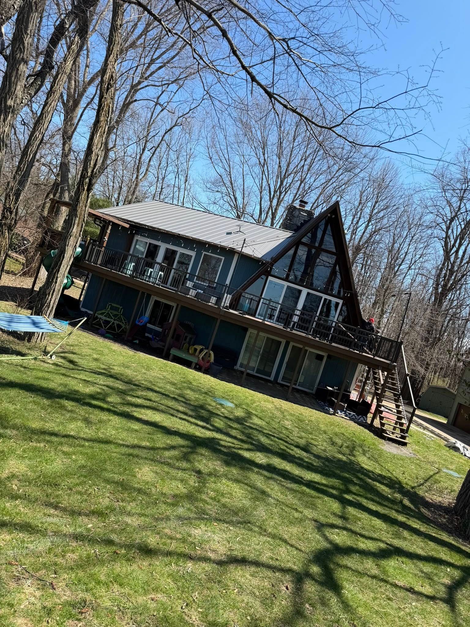 A black-and-white A-frame house with a porch on a grassy slope under leafless trees in bright sunlight