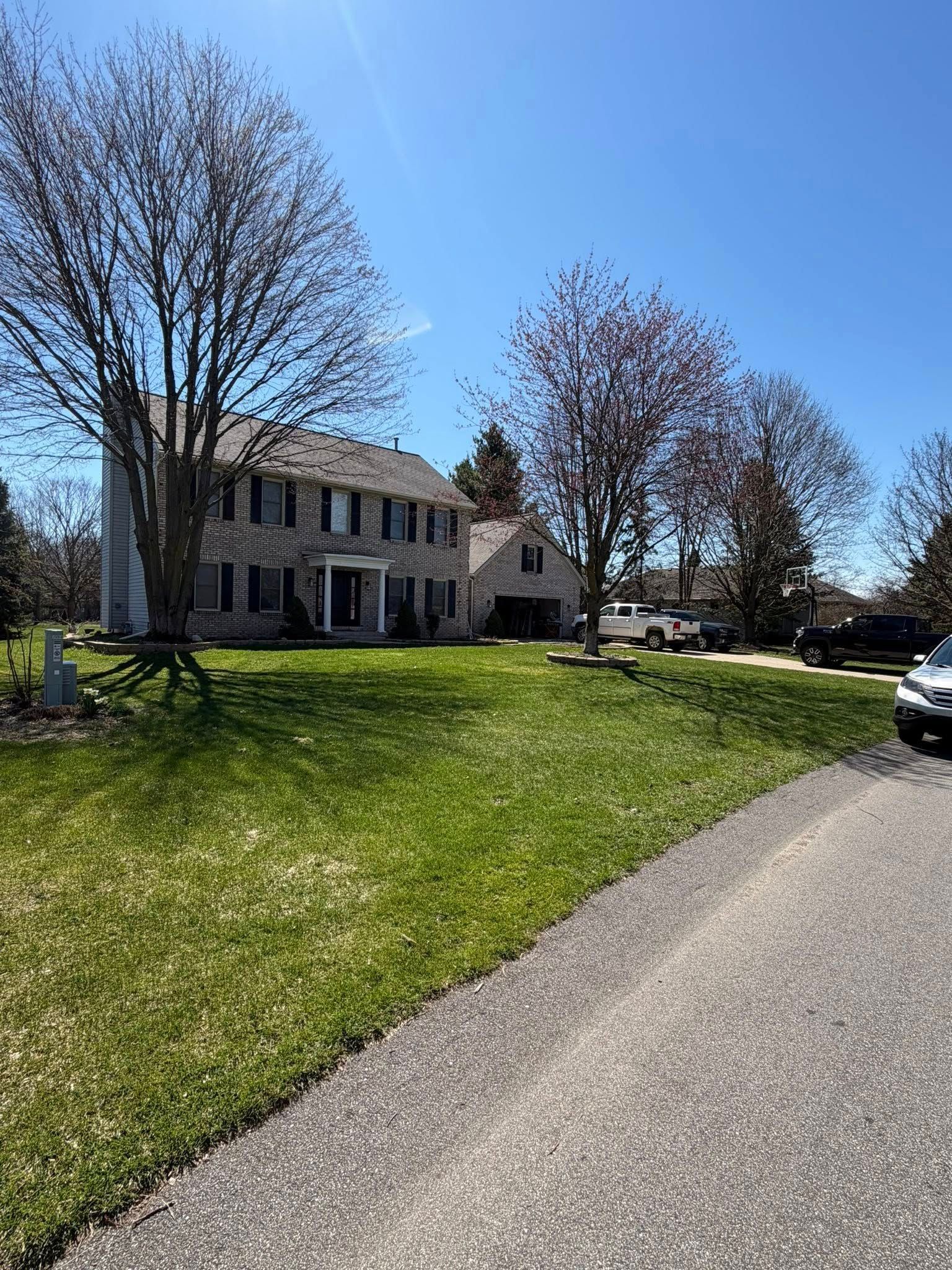 Two-story house with white siding and dark shutters beside a sunlit lawn and driveway under clear blue sky