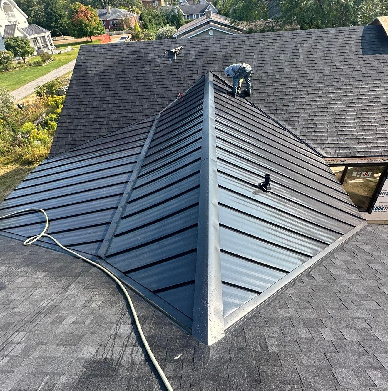 Roofing worker inspecting a dark gray shingle roof with a metal ridge on a house.