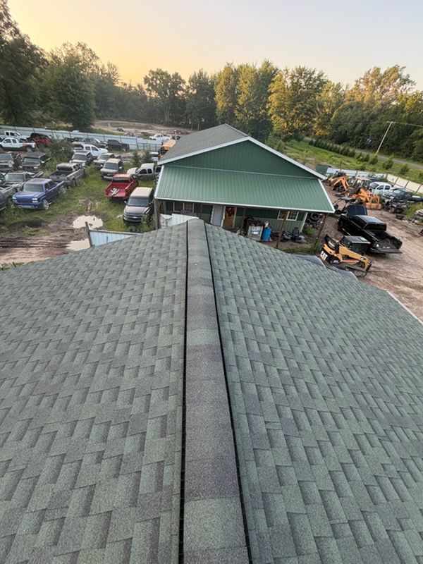 View from a shingled roof over a green-roofed building, parking area, and trees at sunset