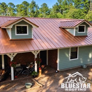 Aerial view of a blue house with a copper metal roof and two dormer windows, front porch below.