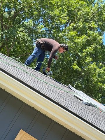 Worker repairing shingles on a sloped roof beside green trees