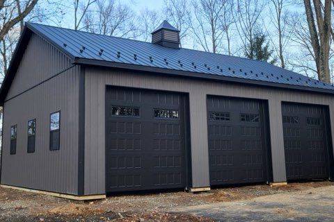 Gray garage building with three black doors and a metal roof, set among bare trees
