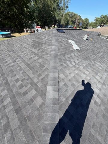 Rooftop with gray shingles, vents, and tree-lined street in the background under bright sunlight.