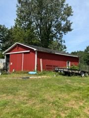 Red barn-style shed with white trim on a grassy lot under a blue sky, with a trailer beside it.
