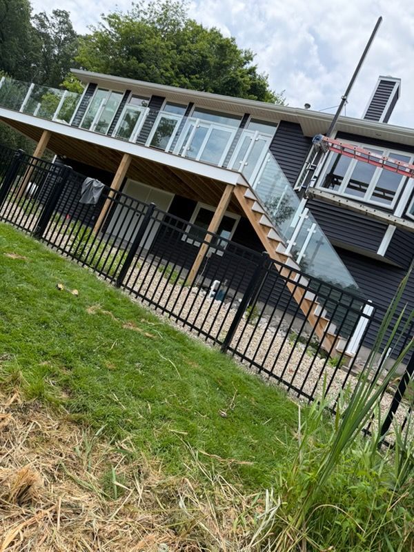 Tilted view of a house deck with glass railing, exterior stairs, and black fence on a grassy slope