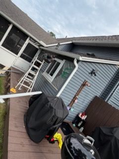 Tilted view of a gray house exterior with a ladder, covered grill, and patio items in the yard.