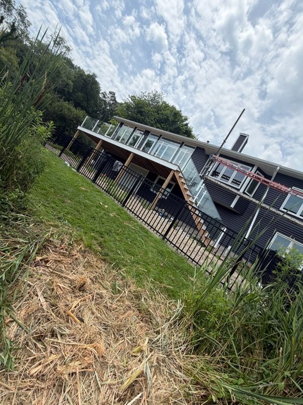 Tilted view of a black riverside building with a deck, seen beyond tall grass and a fence under a cloudy sky