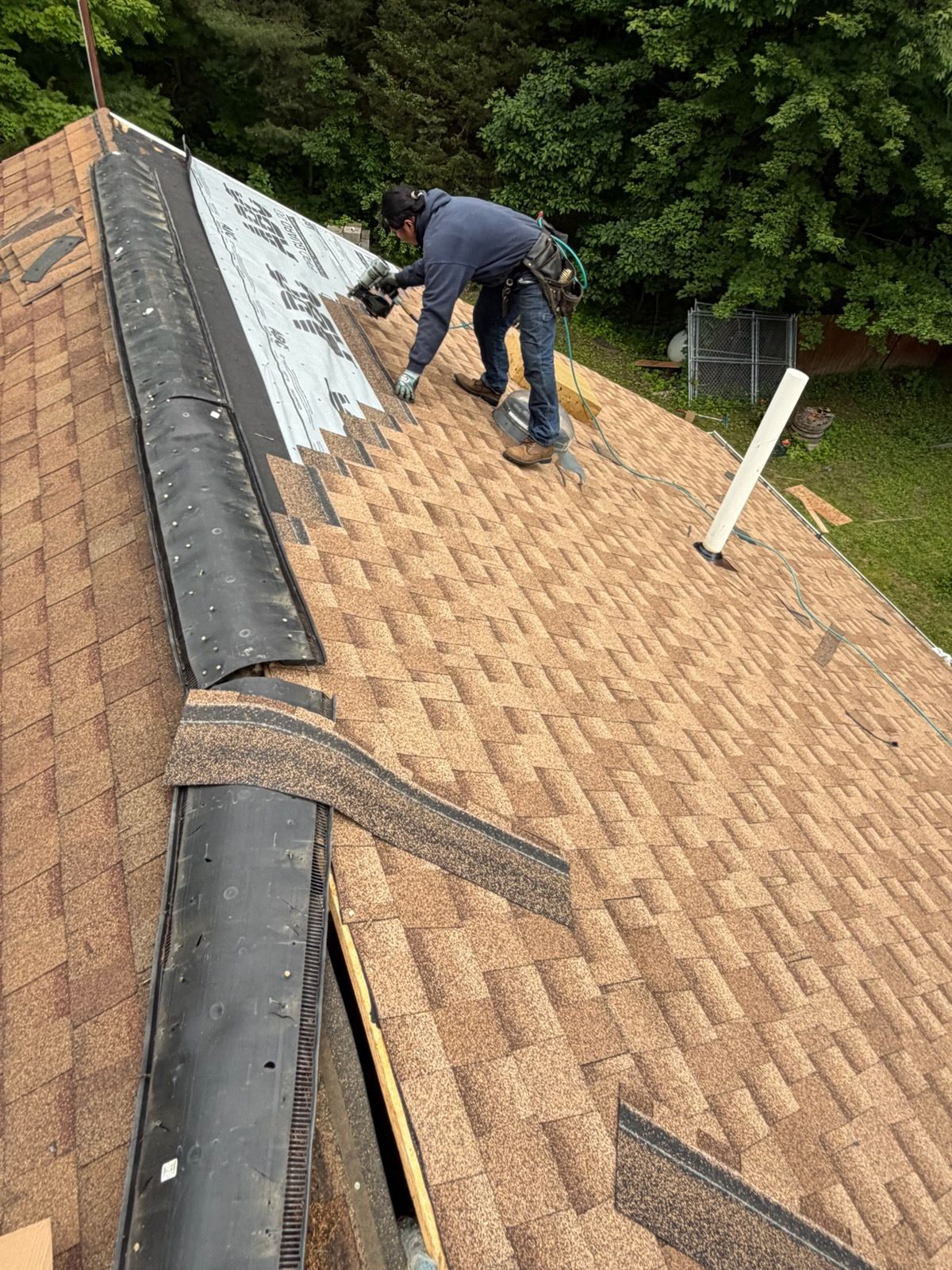 Worker repairing shingles on a steep roof with exposed underlayment and a ladder nearby.