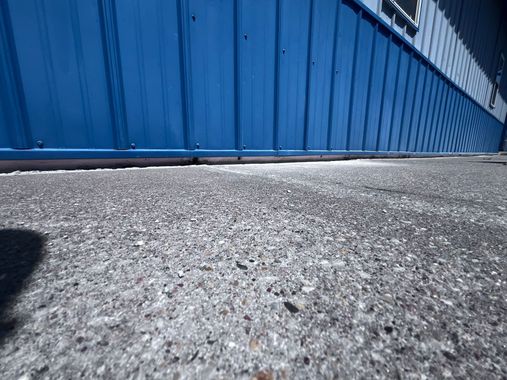Low-angle view of a blue corrugated metal wall beside rough gray pavement
