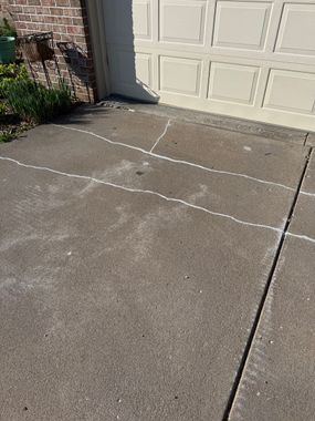Concrete driveway with white chalk markings leading to a garage door, beside a brick wall and plants.