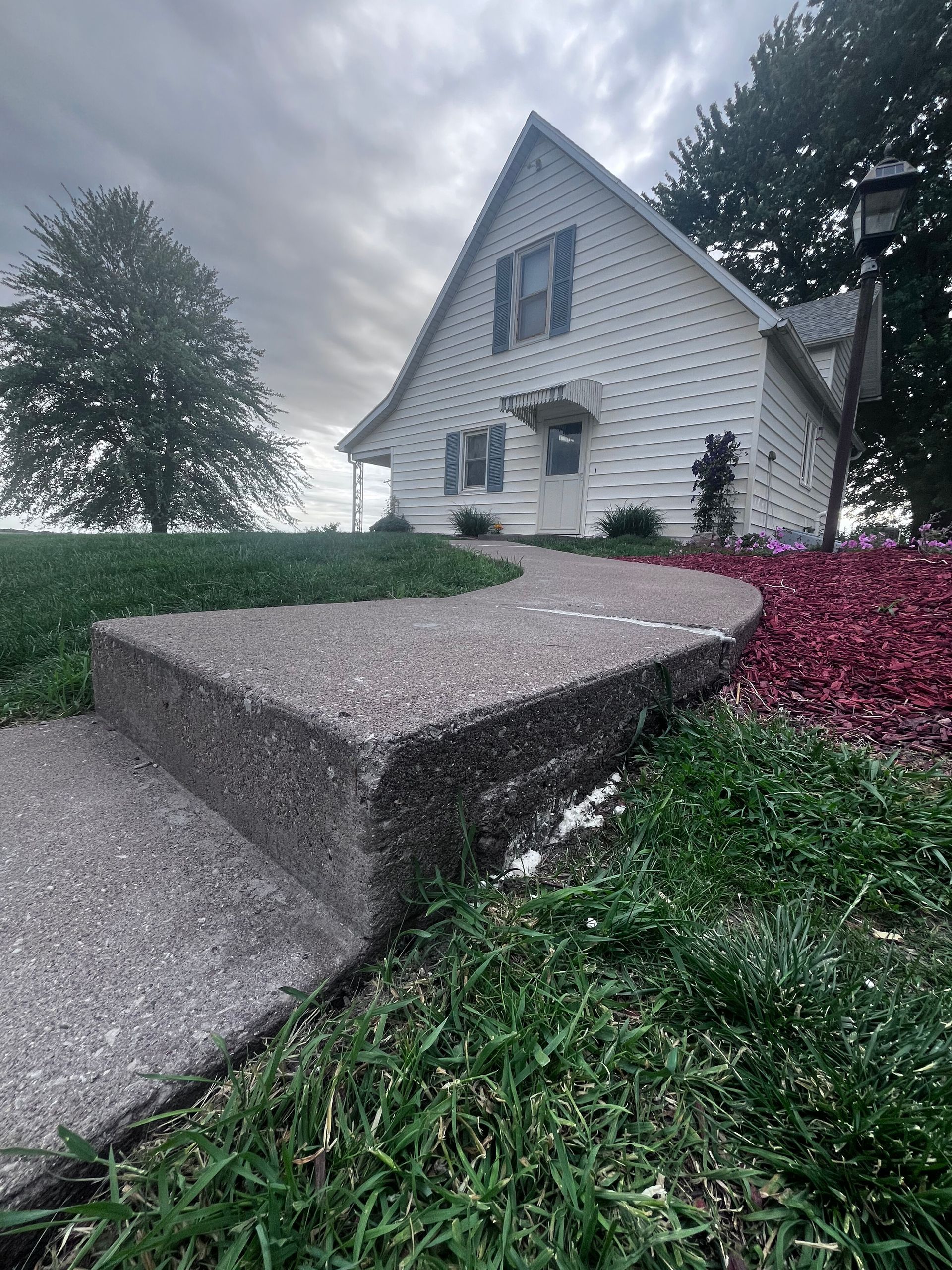White house with a sloped concrete walkway, green lawn, and red shrubs under a cloudy sky.