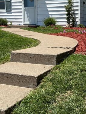 Curved concrete walkway with a raised step leading to a white house entrance and landscaped lawn.