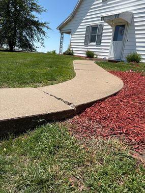 Curved concrete walkway beside a white house, bordered by green lawn and red mulch under bright sun