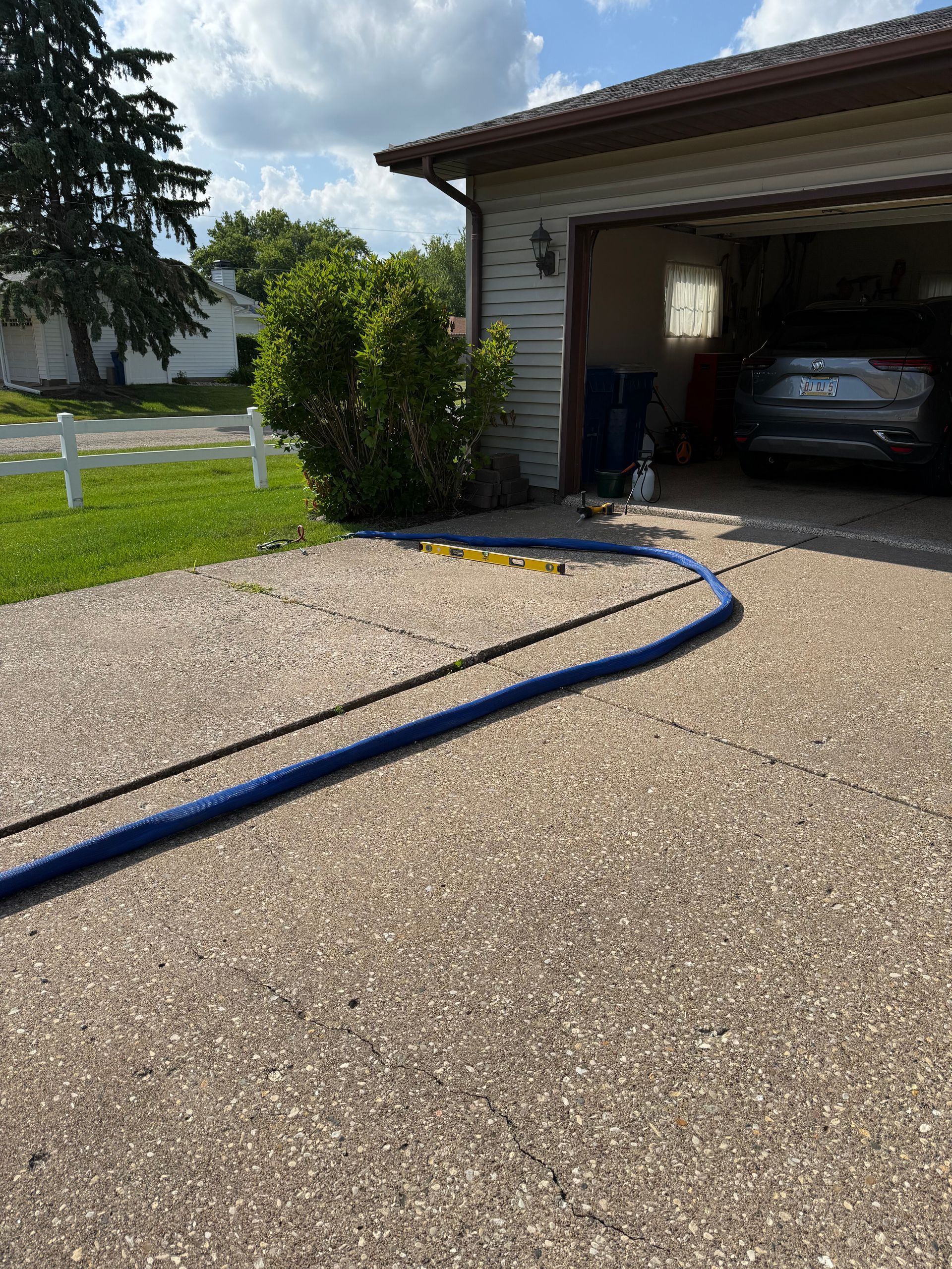 Blue hose on gravel driveway leading to an open garage with a car inside, beside a lawn and tree.
