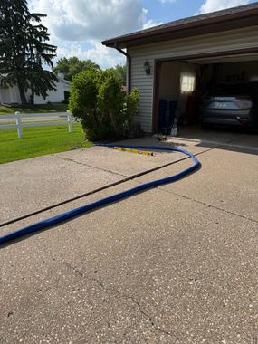 Gravel driveway with a blue hose leading to an open garage and parked car, beside a lawn and tree.