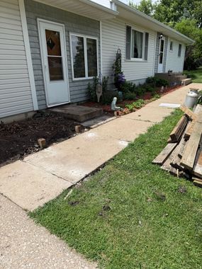 Concrete walkway beside a light-gray house with windows, flower bed, and stacked boards on a grassy lawn