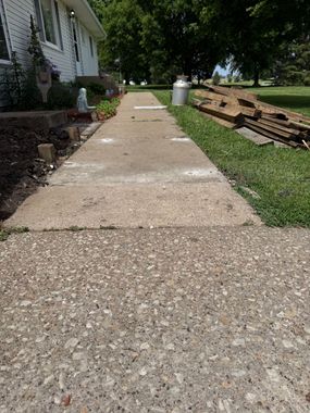 Front sidewalk beside a house, with stacked wood on the right and grass along the edge.