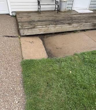 Weathered wooden porch with a cracked concrete step beside a green lawn and gravel driveway.