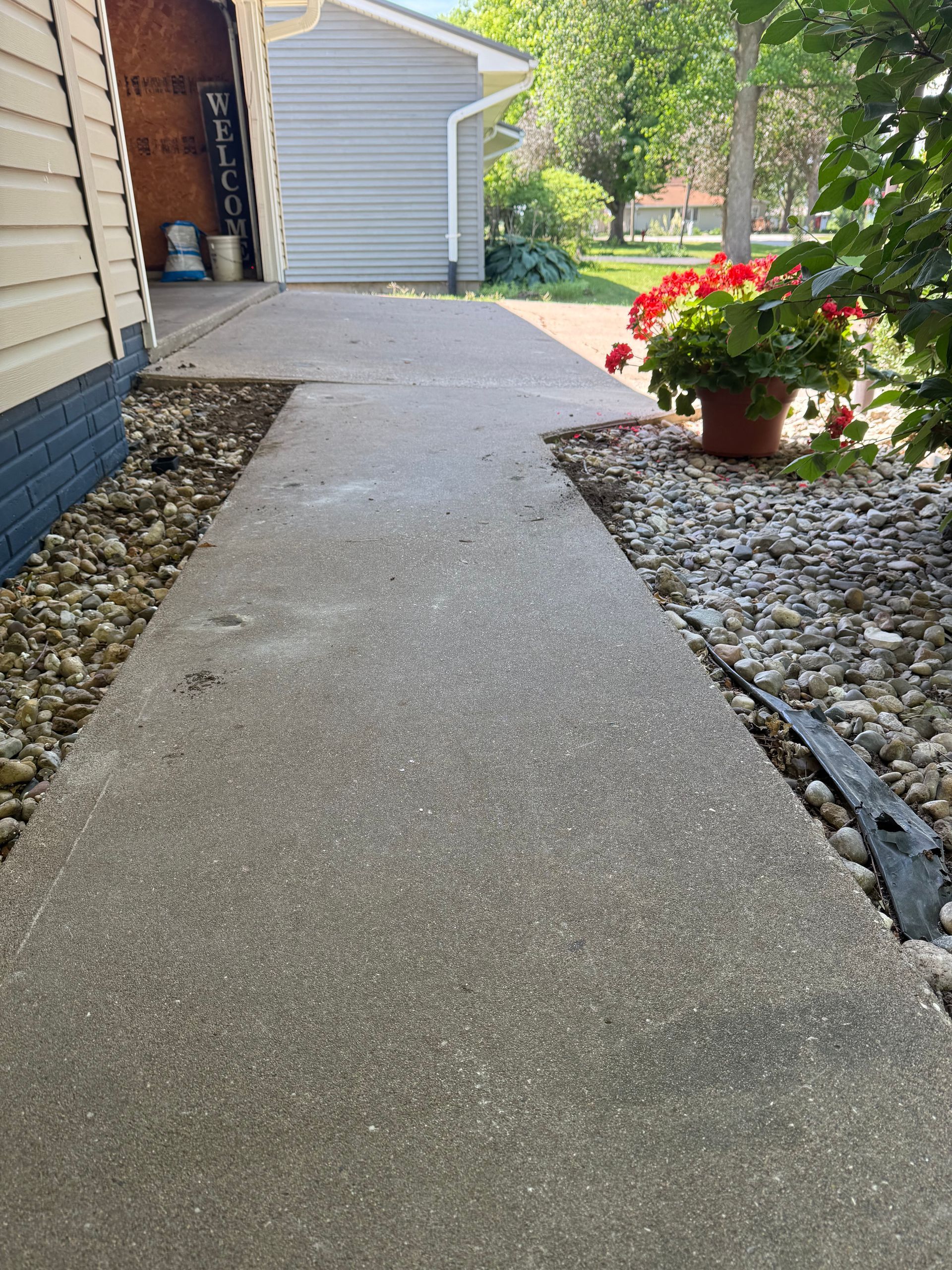 Concrete walkway beside a house, with gravel edging and a red flowering potted plant near the garage.