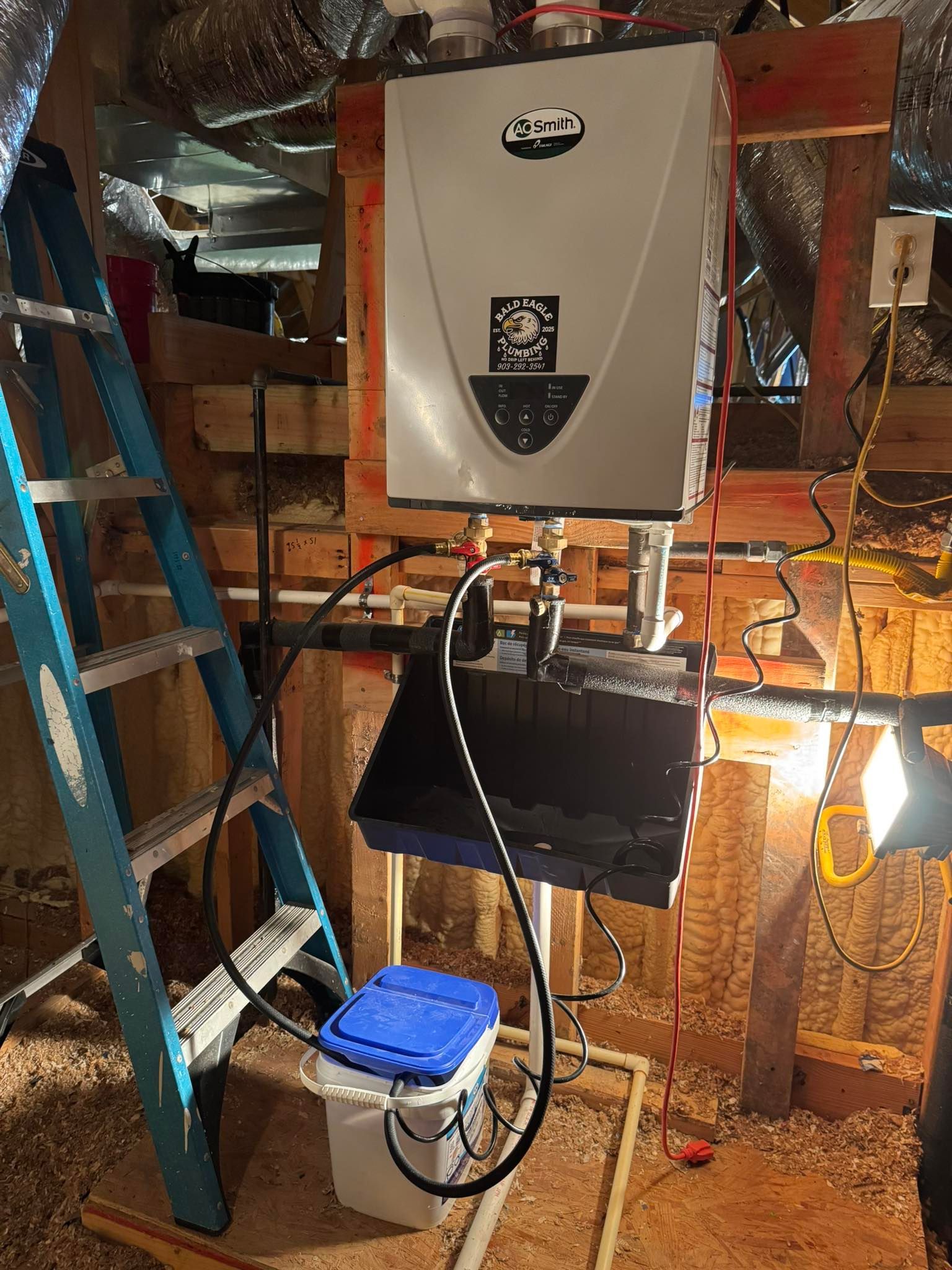 A wall-mounted tankless water heater in an attic, with a pump and blue-lidded bucket beneath it for maintenance.