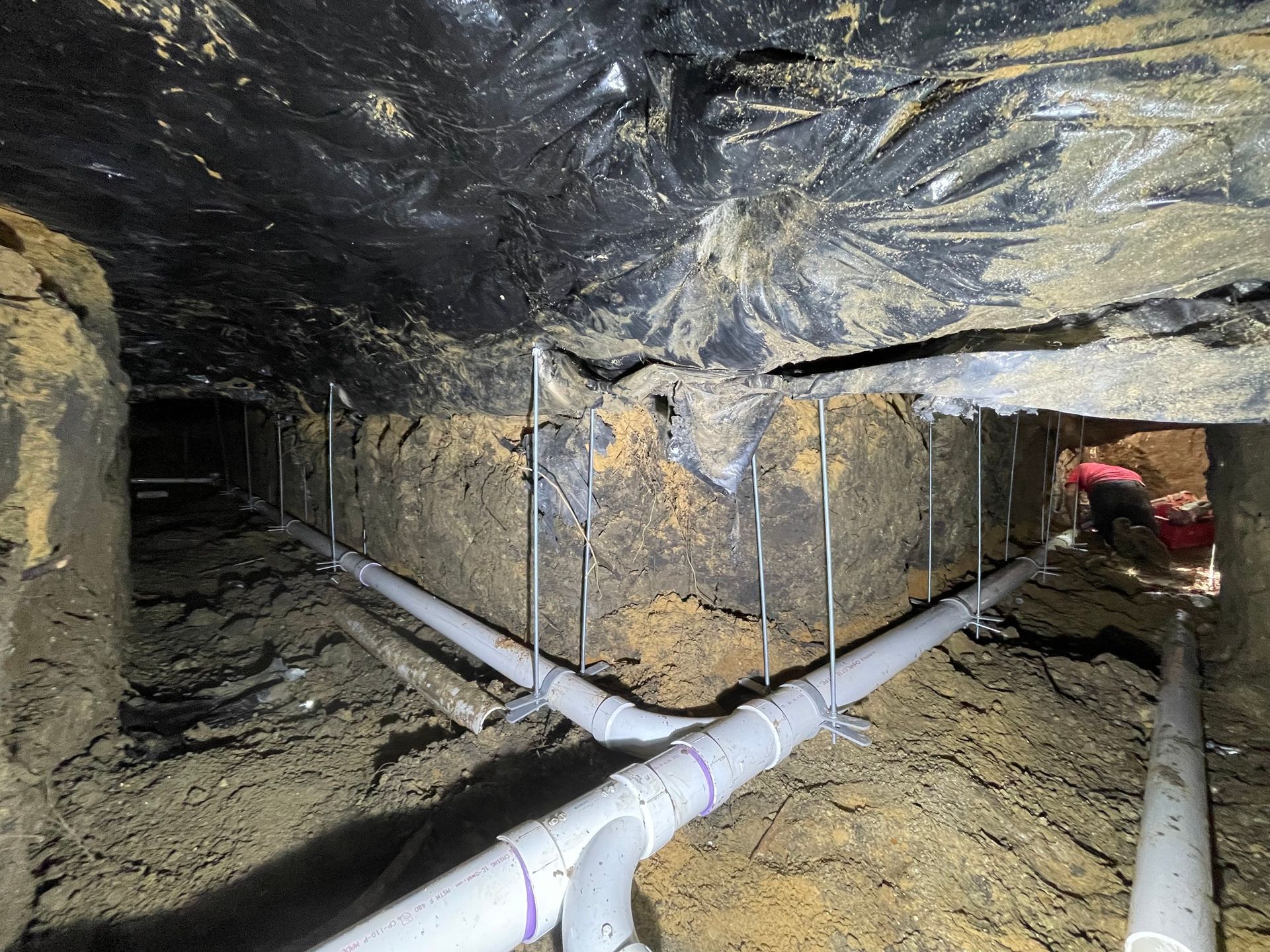 New grey PVC plumbing pipes installed under a house foundation with vertical metal supports in a crawlspace.
