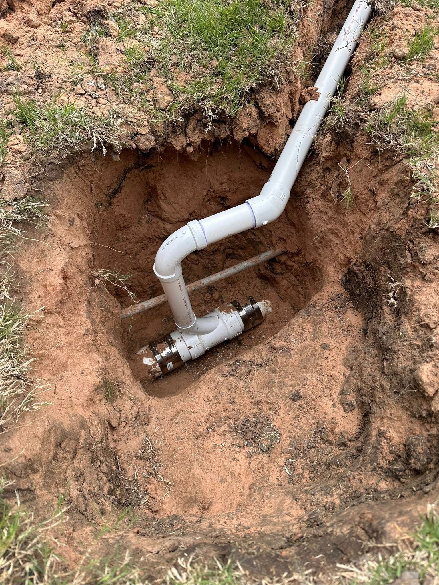 A white PVC pipe assembly with a T-junction connector installed in an excavated dirt trench.