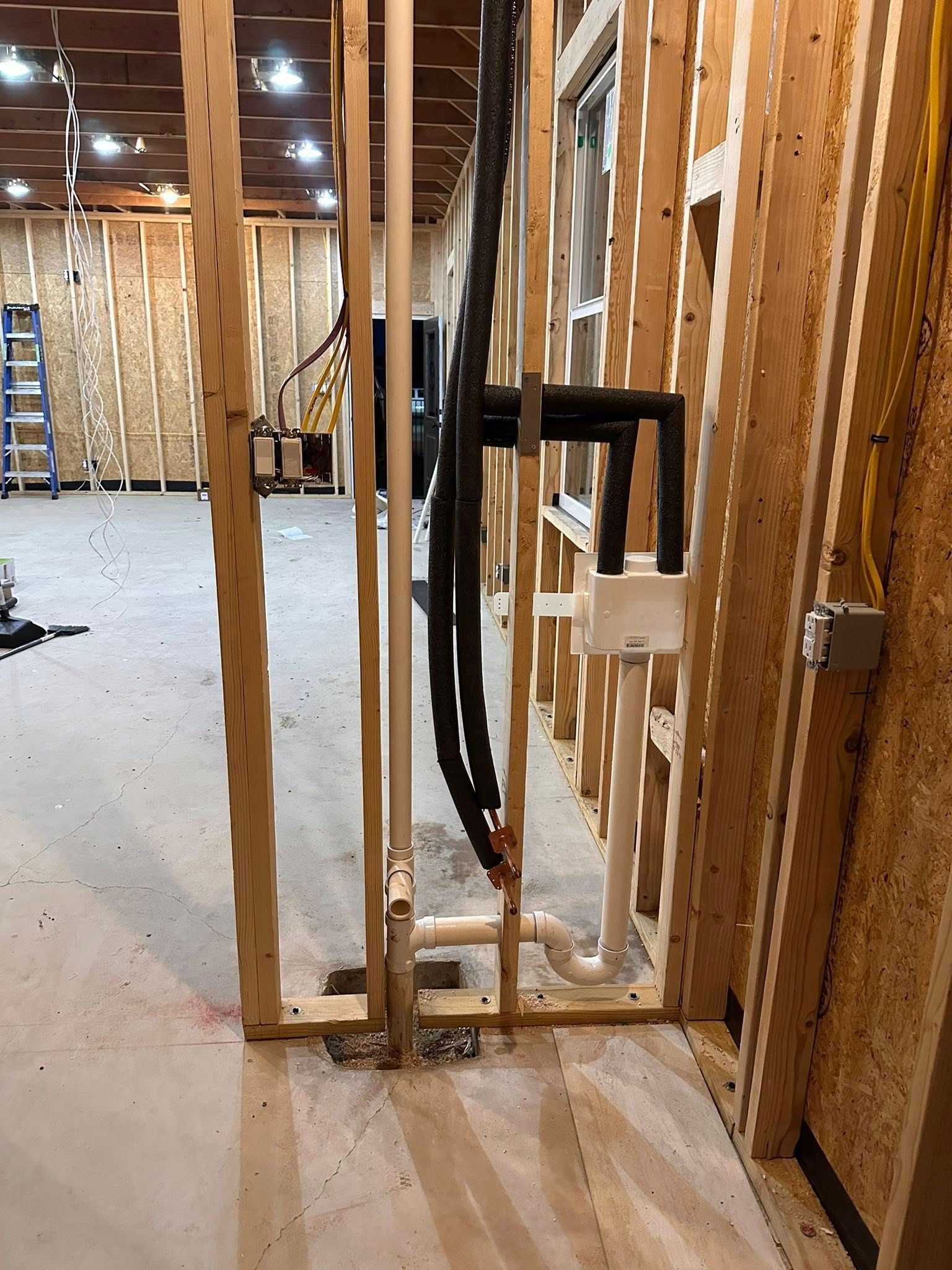 Interior construction site showing wall studs, insulated piping, and plumbing infrastructure in an open framed room.