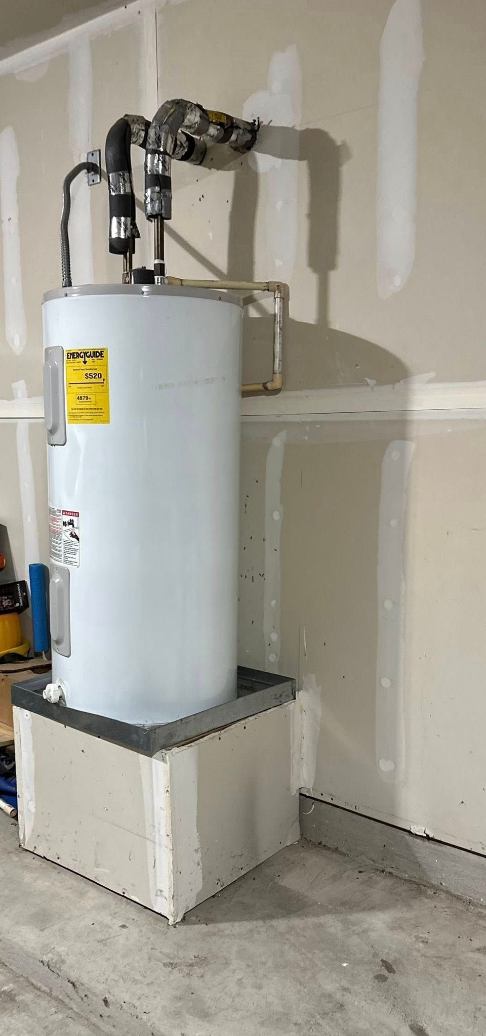 A white water heater stands on a raised drywall platform in a garage, connected to plumbing lines on the wall.
