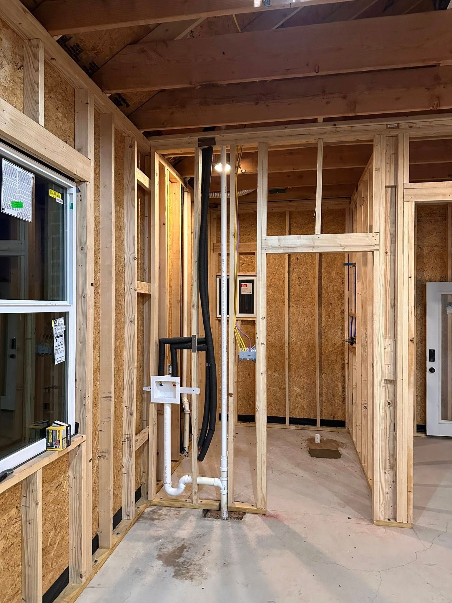 Interior construction frame showing wooden studs, an open doorway, and installed plumbing pipes for a laundry area.