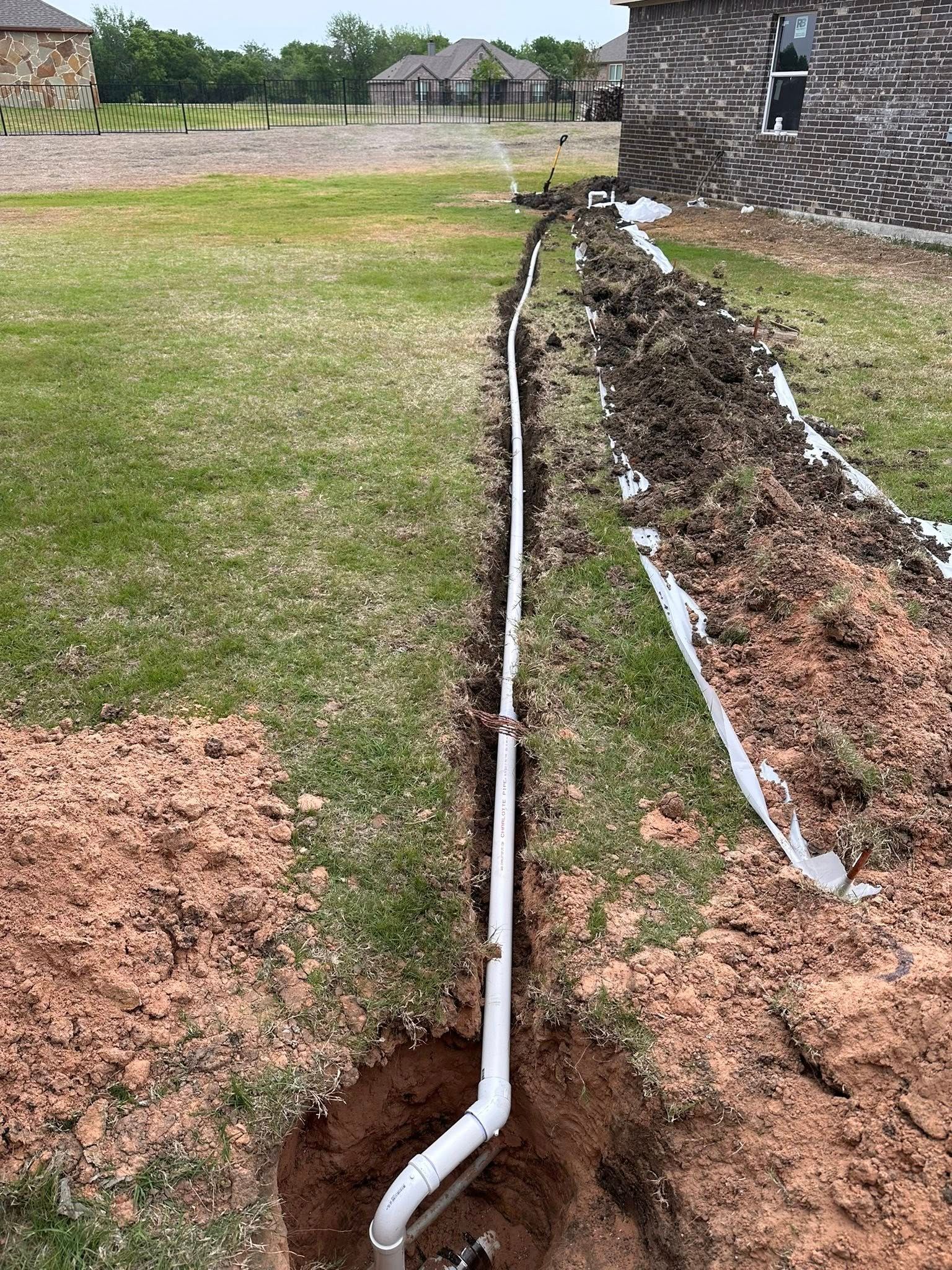 A white PVC pipe runs through a trench in a grassy backyard alongside the brick wall of a house.