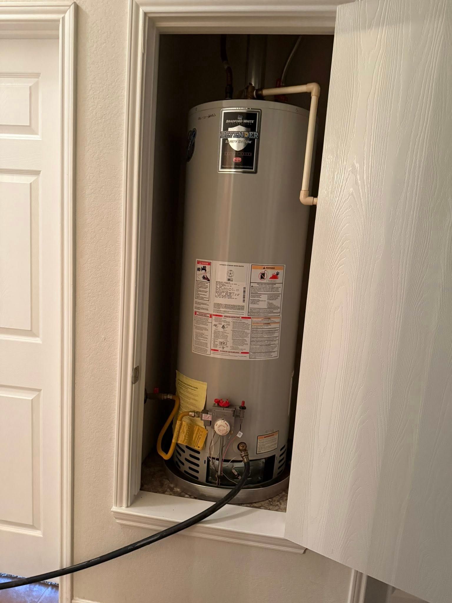 A gray residential water heater installed in a recessed closet, connected to pipes and a flexible gas supply line.