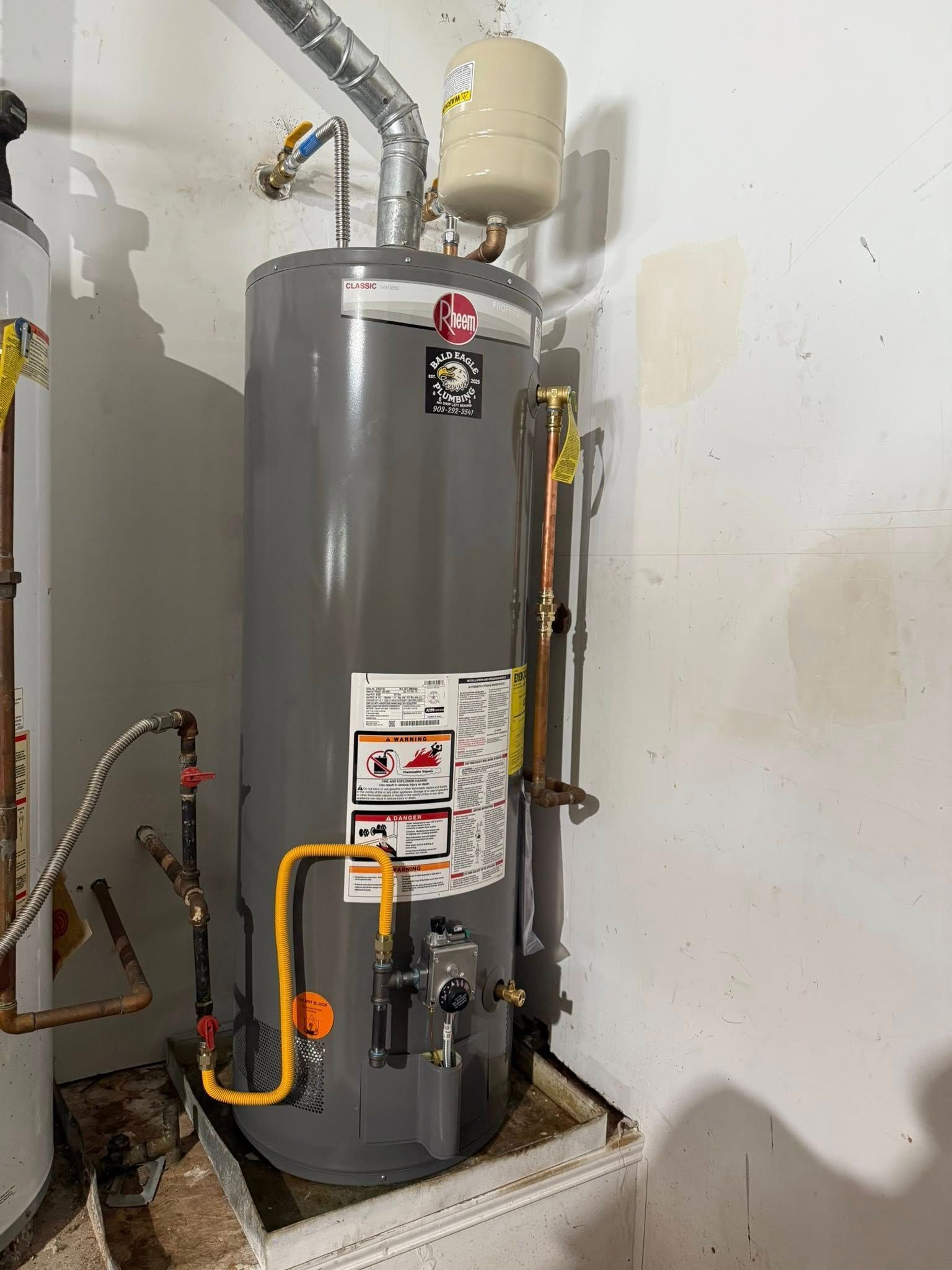 A gray gas water heater sits in a utility closet with an expansion tank, venting, and plumbing connections attached.