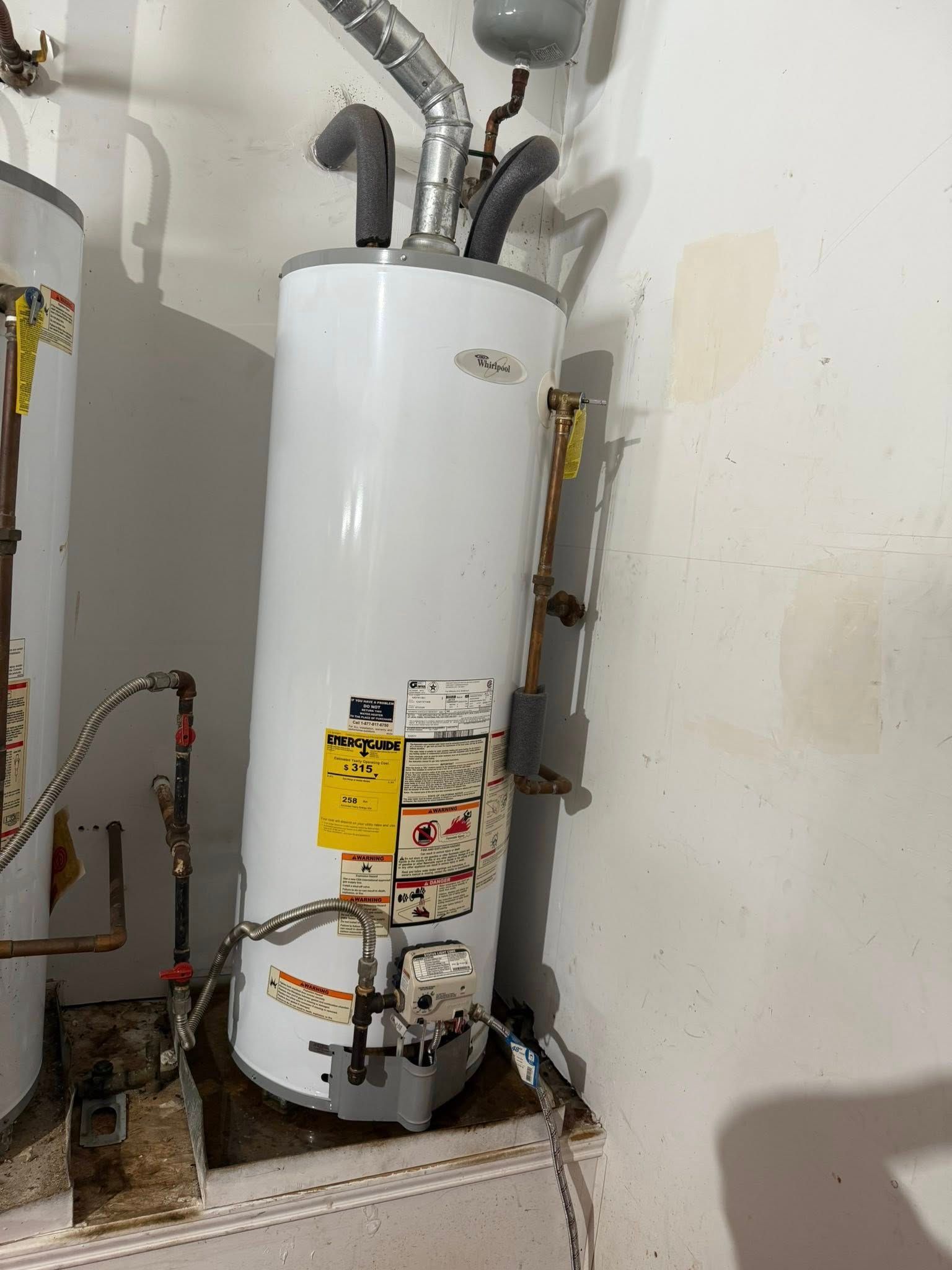 A white gas water heater with plumbing connections and a yellow information label, situated against a beige wall.
