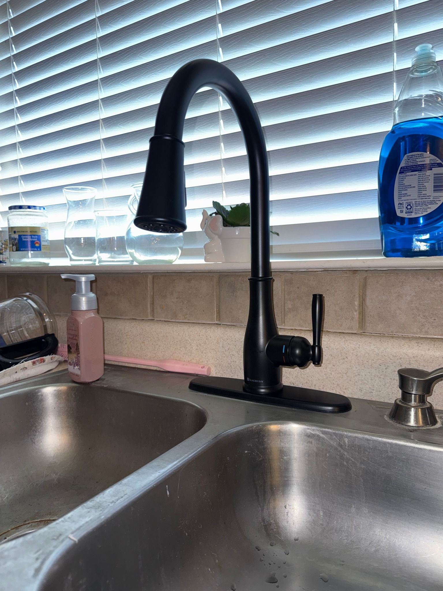 A matte black pull-down kitchen faucet centered between double stainless steel sinks in front of a window with blinds.
