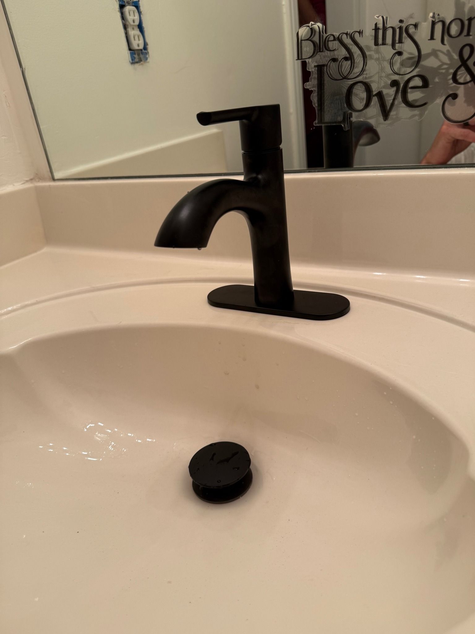 A matte black bathroom faucet and sink drain in a white basin with a bathroom mirror in the background.