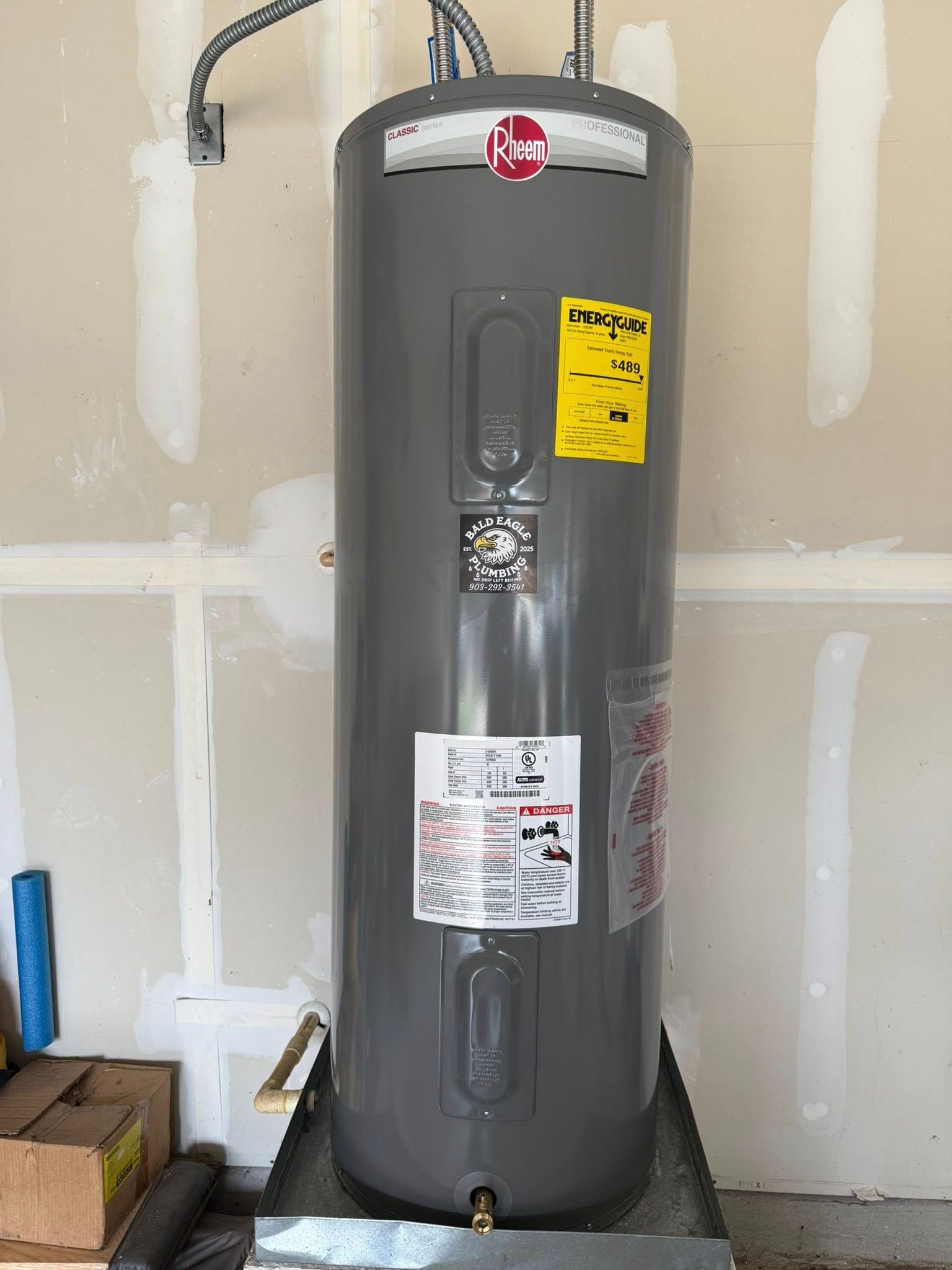 A gray Rheem electric water heater stands upright in a drain pan against an unfinished drywall wall in a garage.