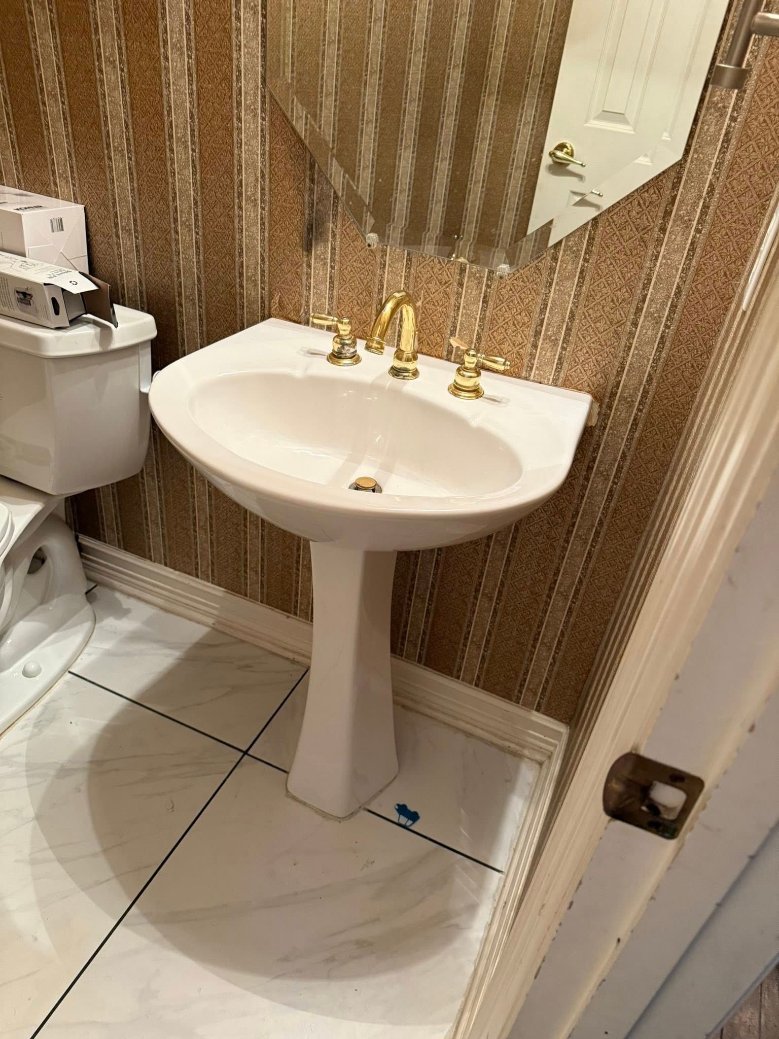 A white pedestal sink with gold fixtures, set against patterned wallpaper and white tiled floors in a small bathroom.