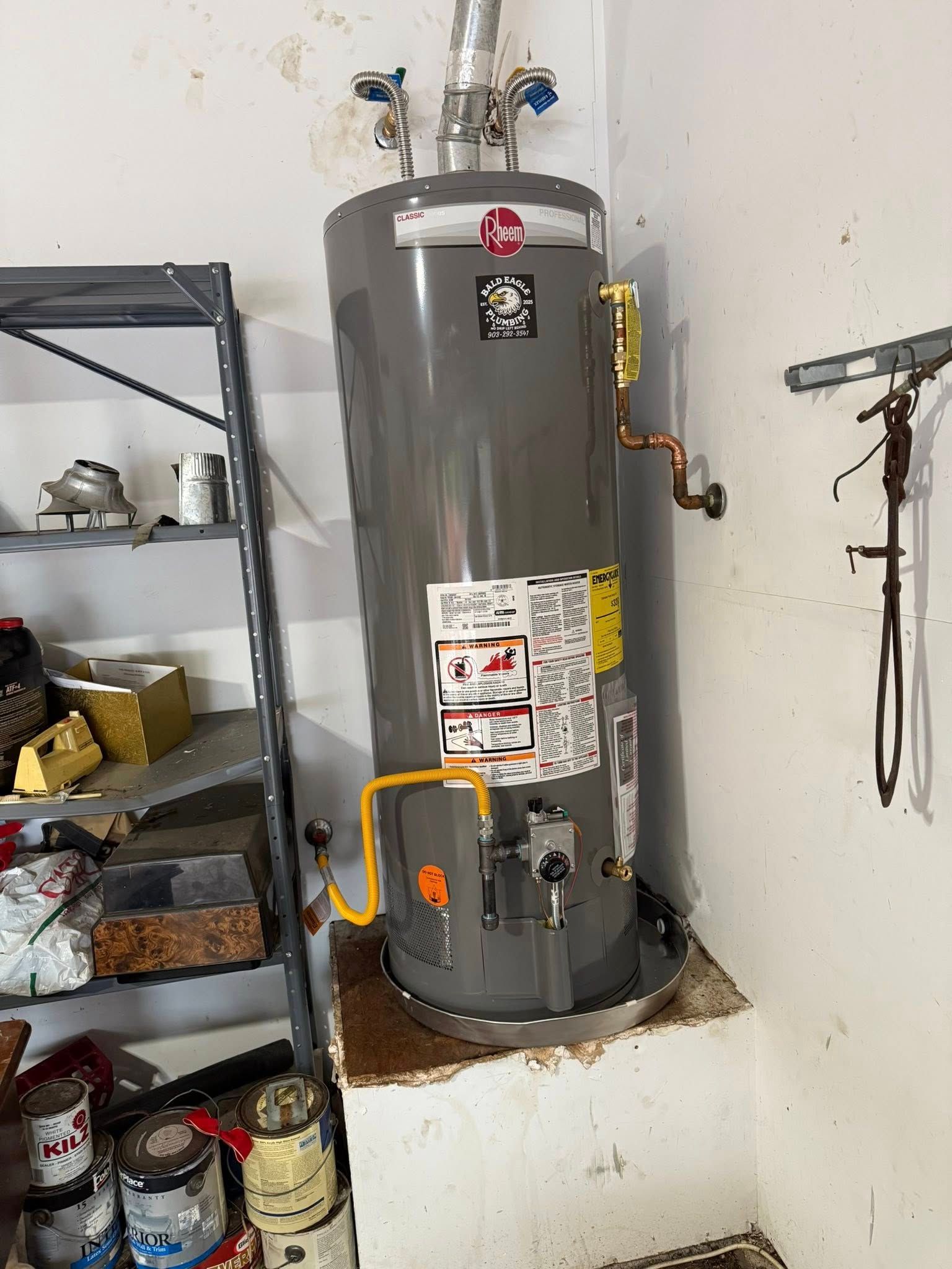 A gray gas water heater sits on a raised platform in a garage next to metal shelving and a tool-hanging rack.