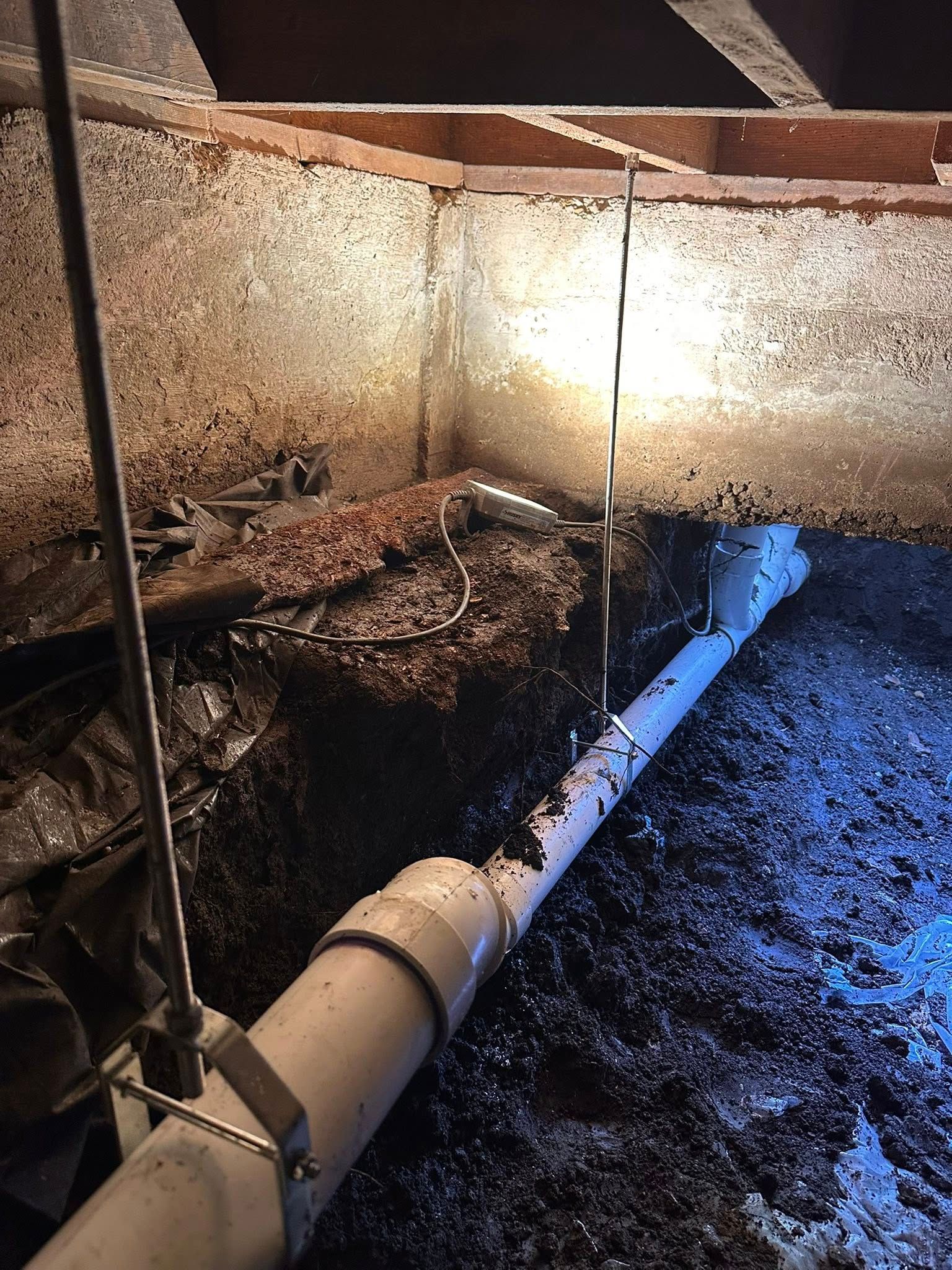 A white PVC pipe runs through a dark crawlspace with loose fill insulation, supported by metal straps from wooden beams.