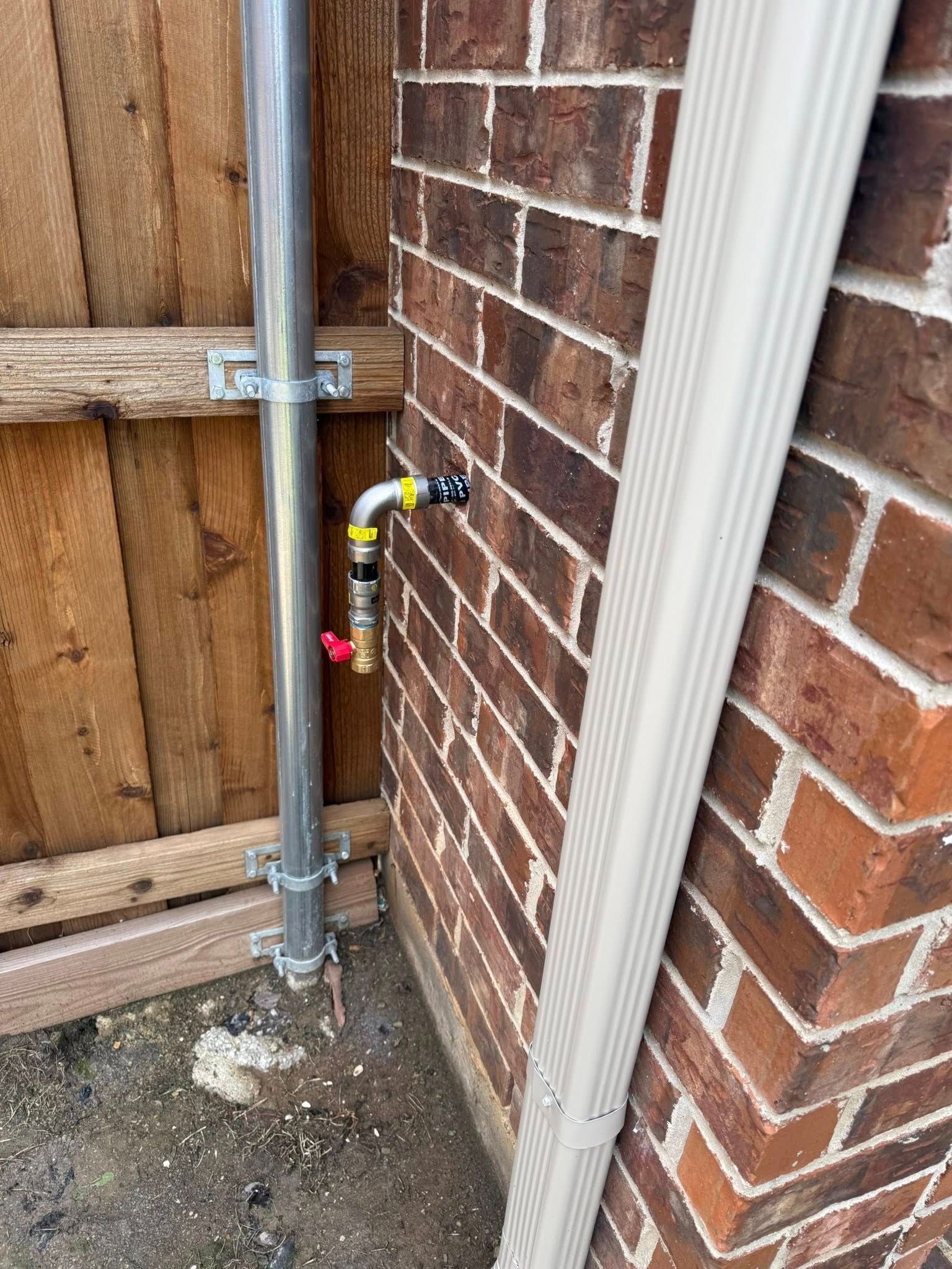 A gas line stub-out with a red shut-off valve protruding from a brick wall next to a metal post and a downspout.