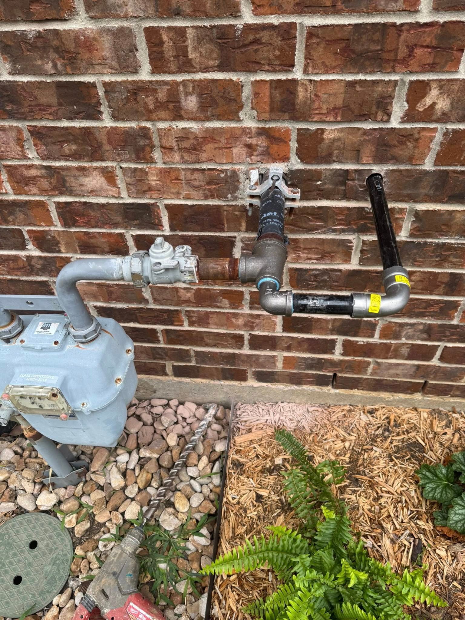 A gas meter and metal piping attached to an exterior brick wall above a garden bed with rocks and a green plant.