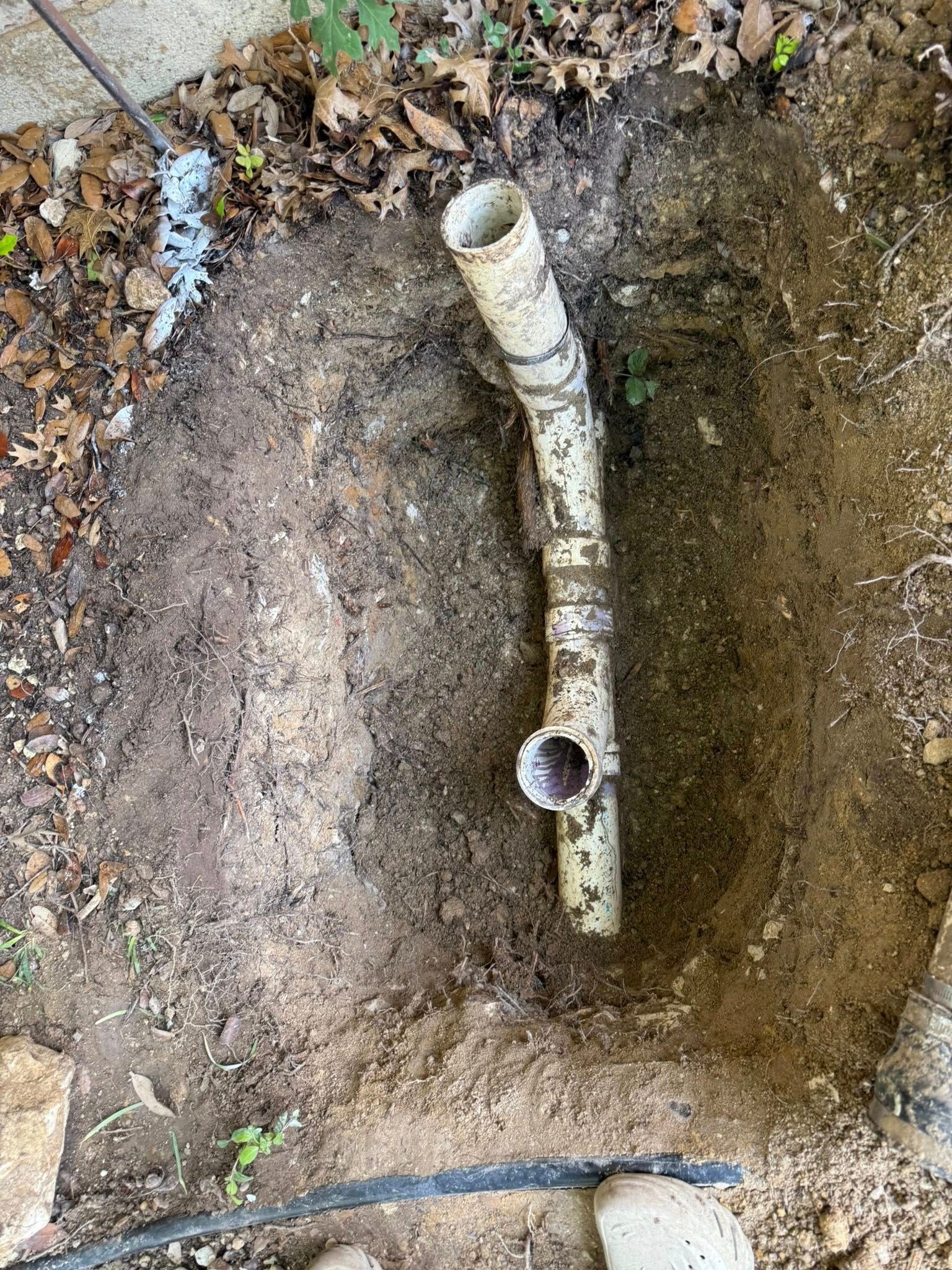 A top-down view of a white PVC drain pipe with a Y-junction fitting buried in a shallow, dirt-filled hole in the ground.