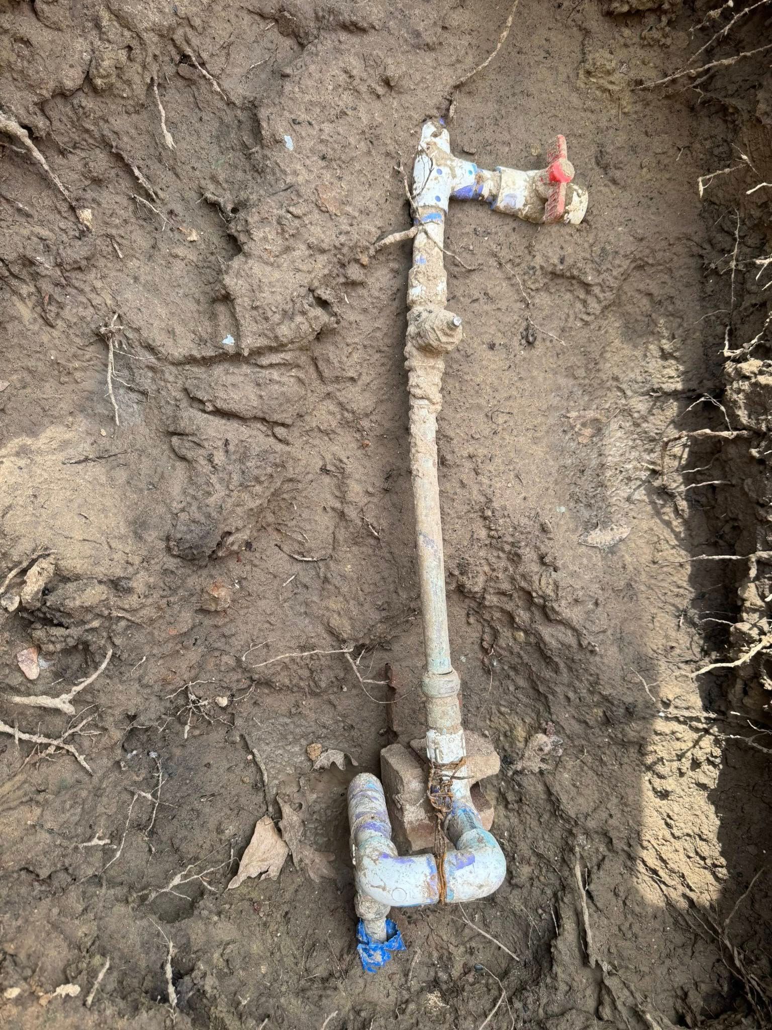 A section of PVC plumbing pipes with a red-handled valve, partially excavated from dark, damp soil.