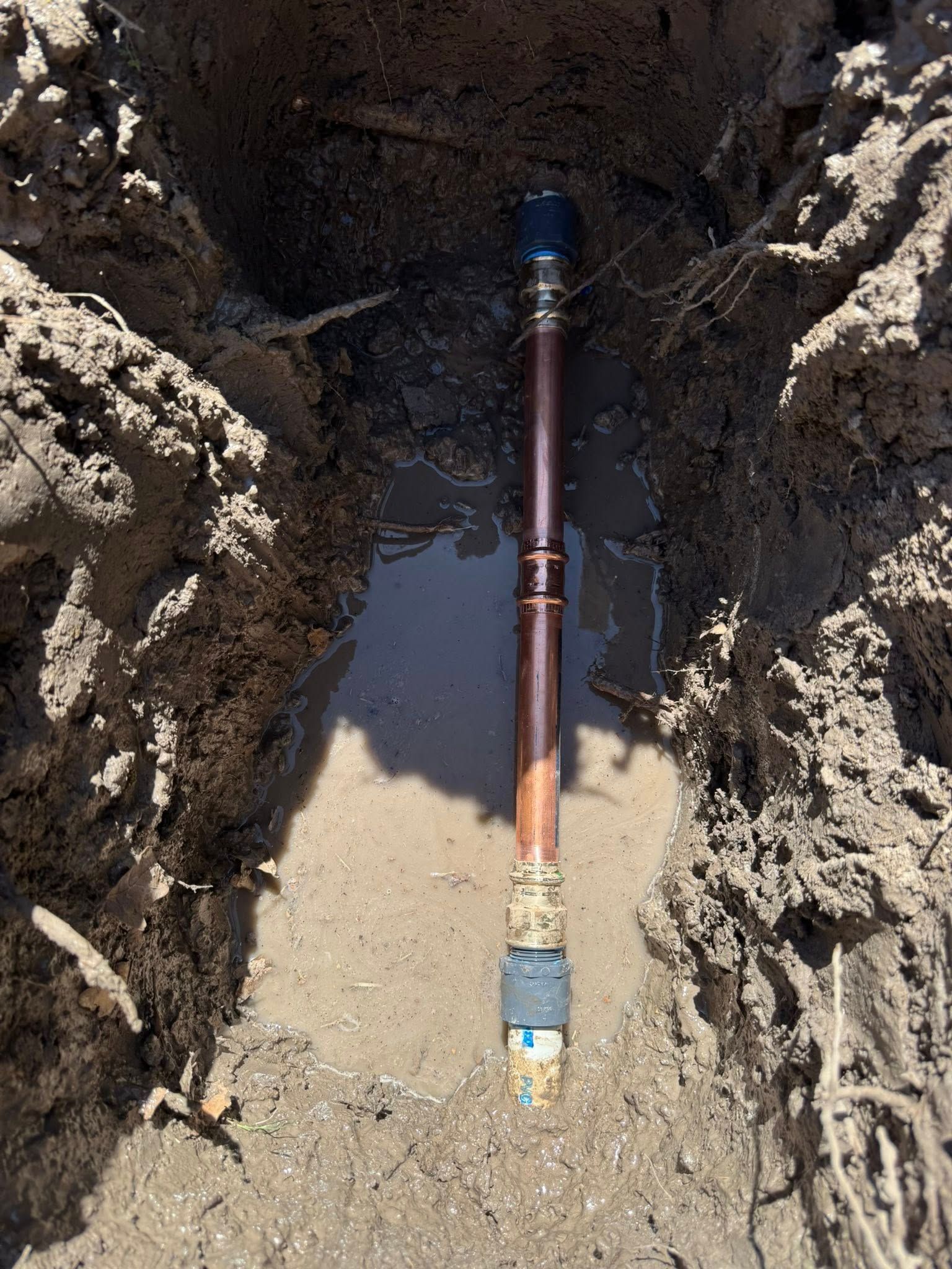 A copper pipe section installed in a narrow, muddy trench in the ground.