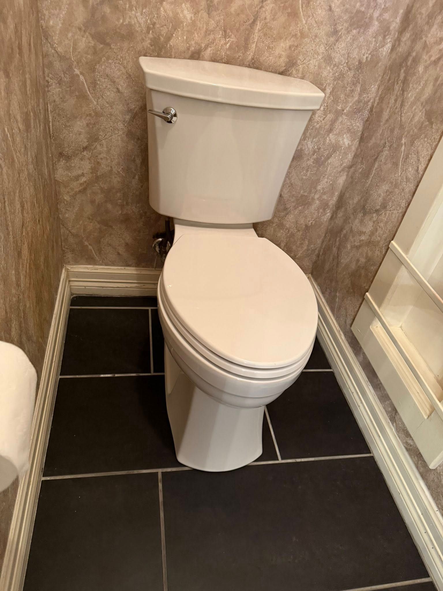 A beige toilet with a closed lid in a bathroom with brown patterned wallpaper and dark floor tiles.