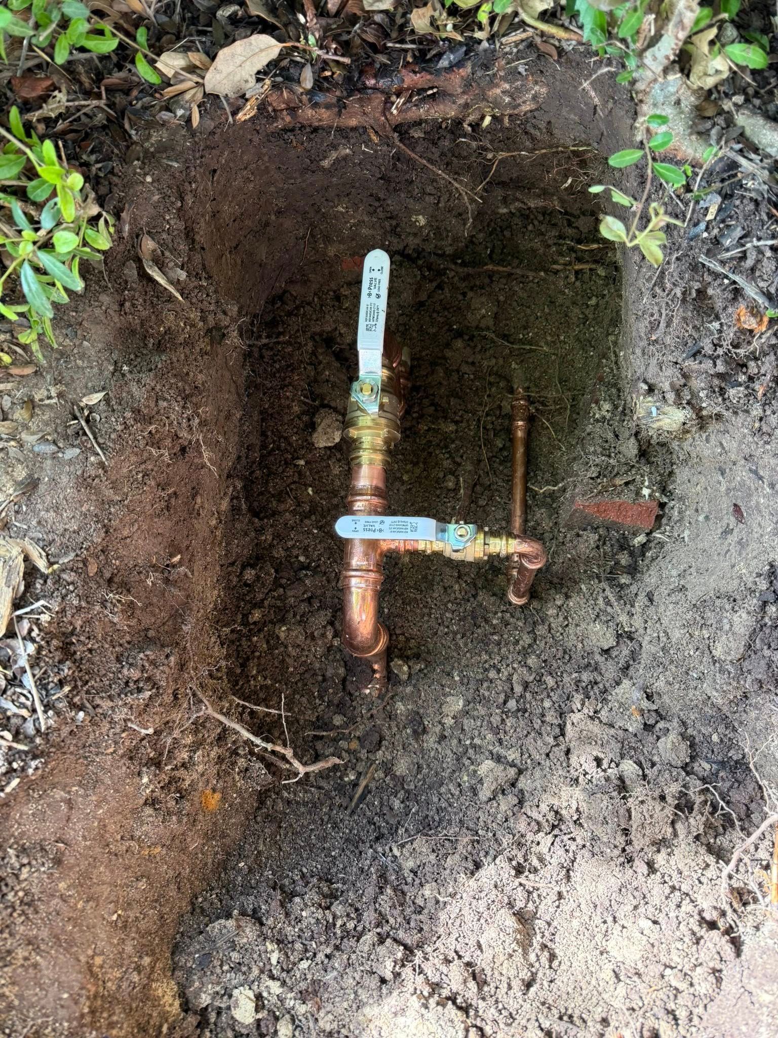 Two copper water valves with blue handles installed in a trench in the dirt.